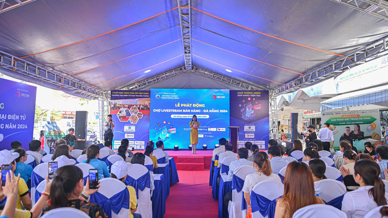 An outdoor event featuring a stage with a presentation, where a speaker stands in front of a large screen displaying event information. Attendees are seated in rows, many with smartphones.