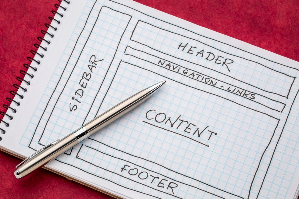 A gold pen lays on top of a notebook of graph paper showing a drawing of a website layout.