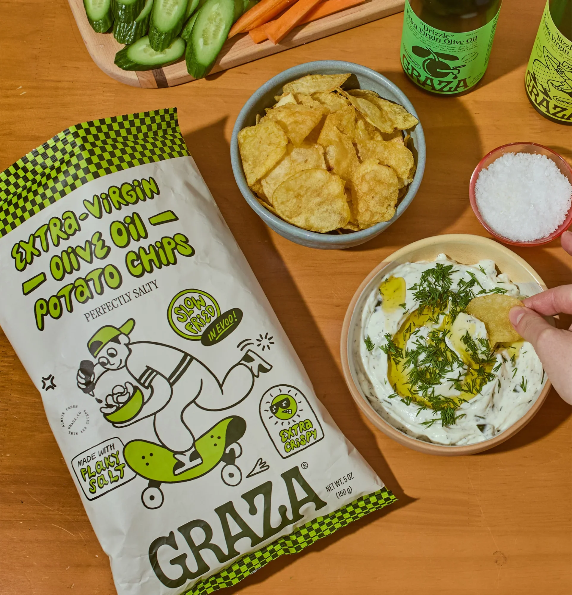 A hand dipping a chip into herby dip next to a bag of Graza olive oil chips on a wooden table.