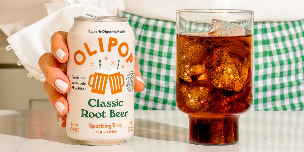 A person holds a can of Olipop's Classic Root Beer next to a tall glass of it, wearing a bright green gingham skirt