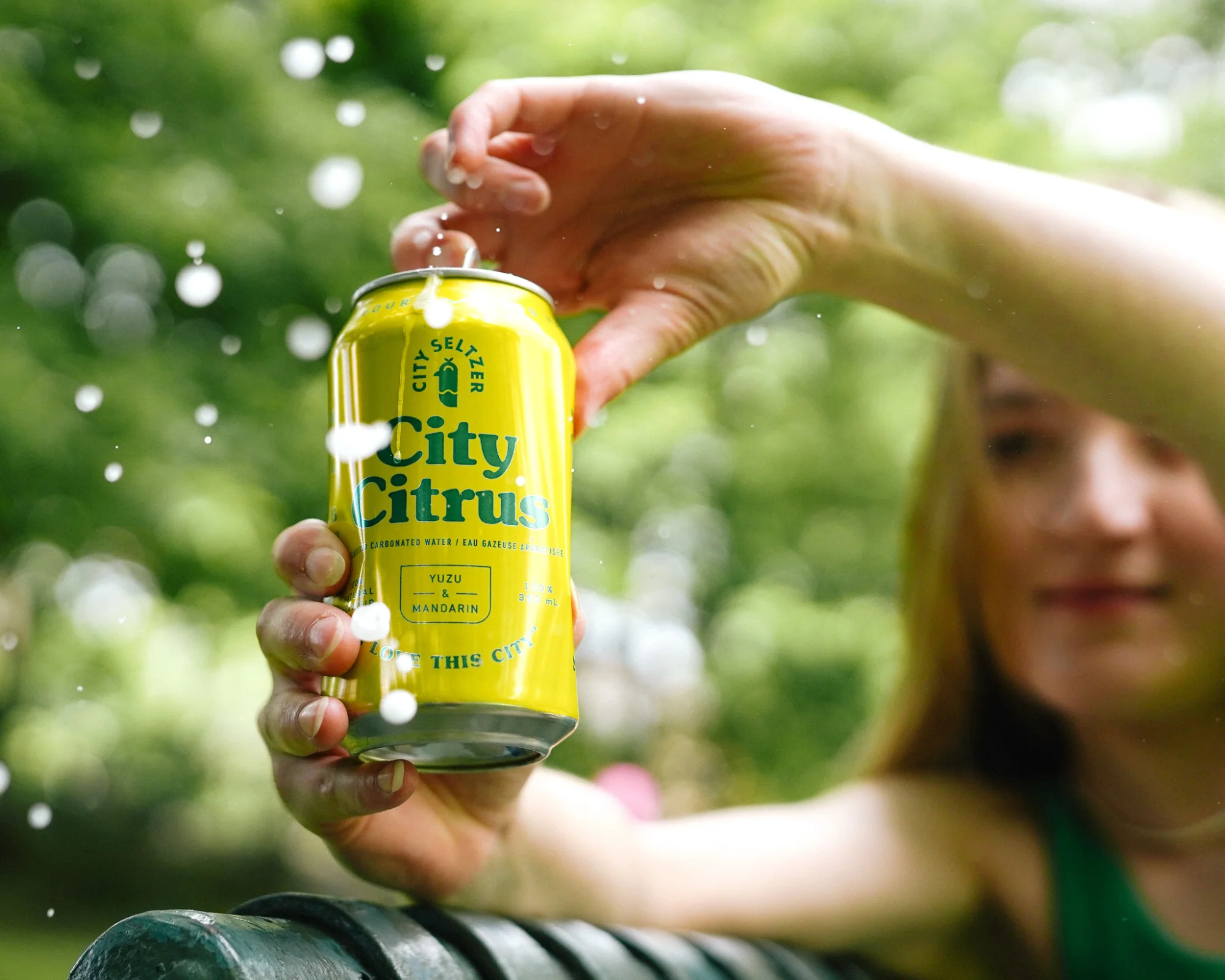 A brightly colored can of City Seltzer is opened by a person sat on a park bench