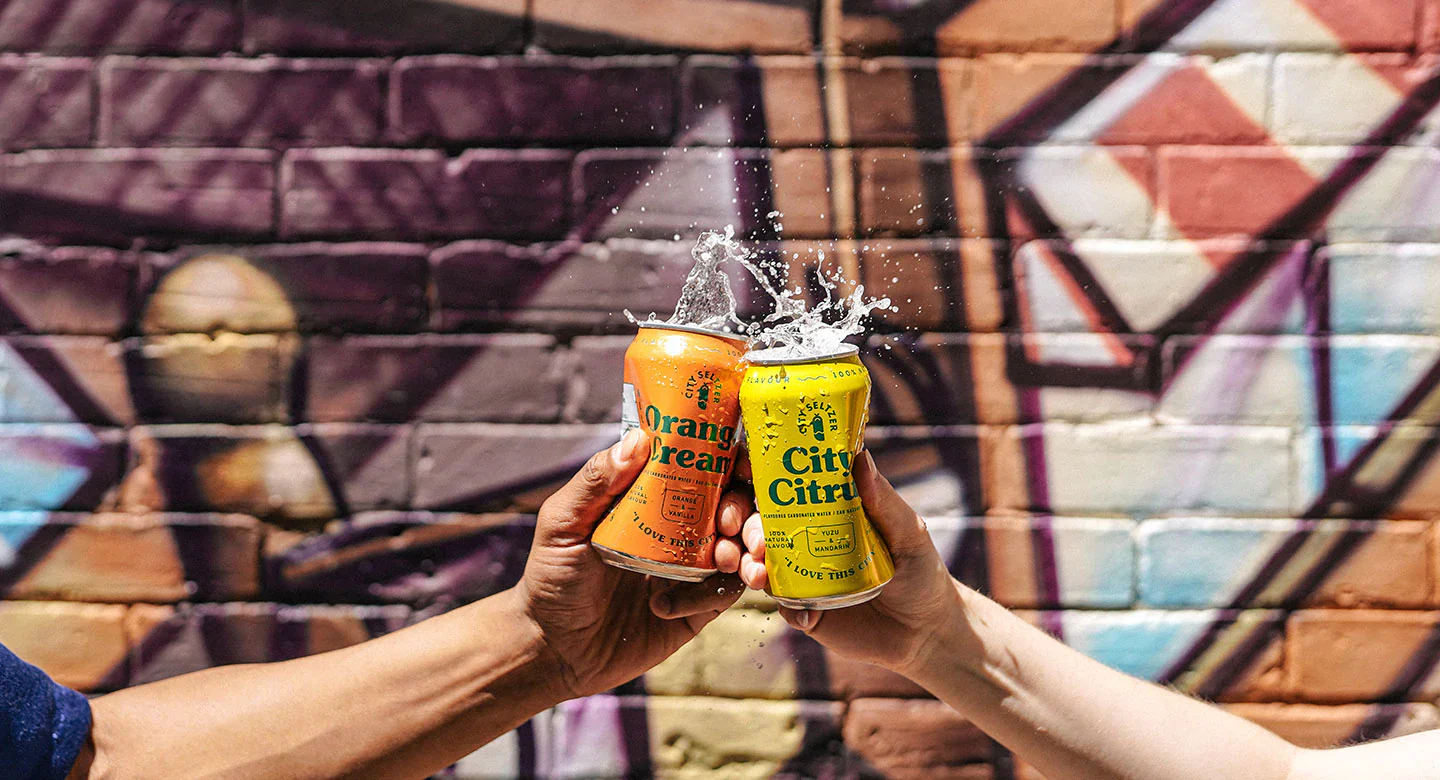 Two people cheering with cans of fizzy pop against a graffitied wall.