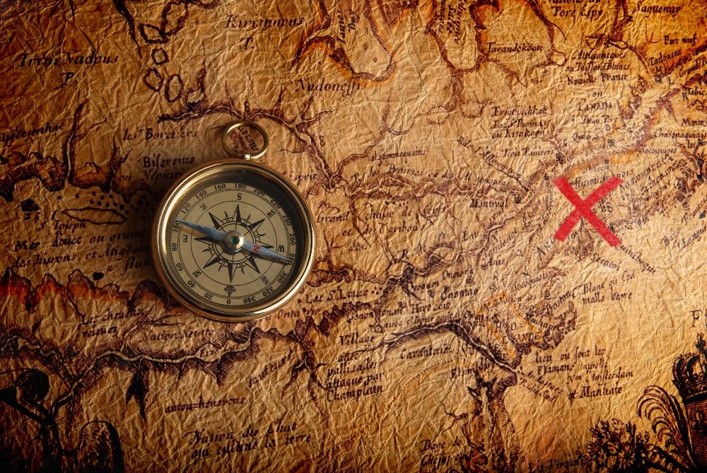A vintage compass resting on an old, textured map marked with a red 'X'. The map features detailed geographical notations and routes in sepia tones, suggesting an ancient or historical theme. The compass is oriented towards the northeast, adding a navigational element to the scene in piece about the Google algorithm leak. 