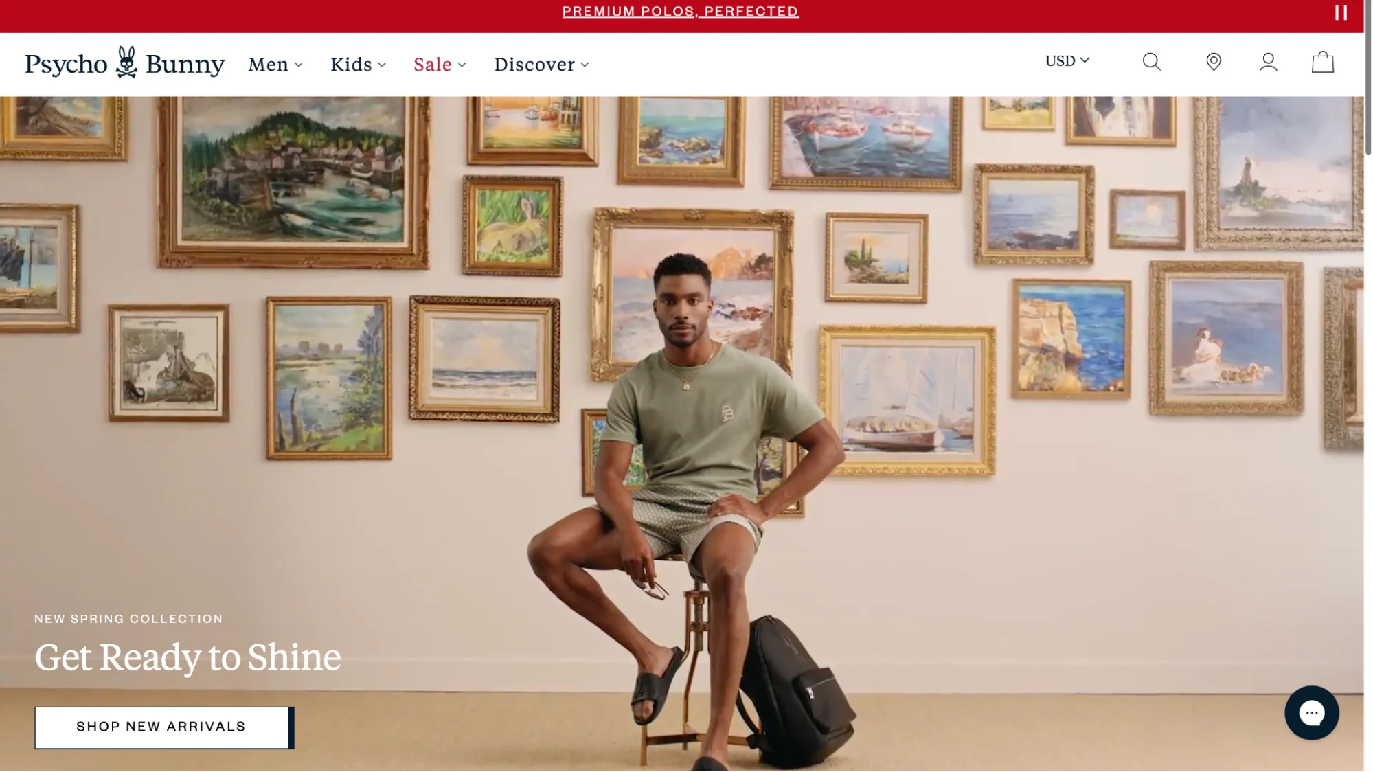 A model sits on a stool, wearing a green shirt and shorts, against a wall adorned with various framed paintings, promoting a new spring collection.