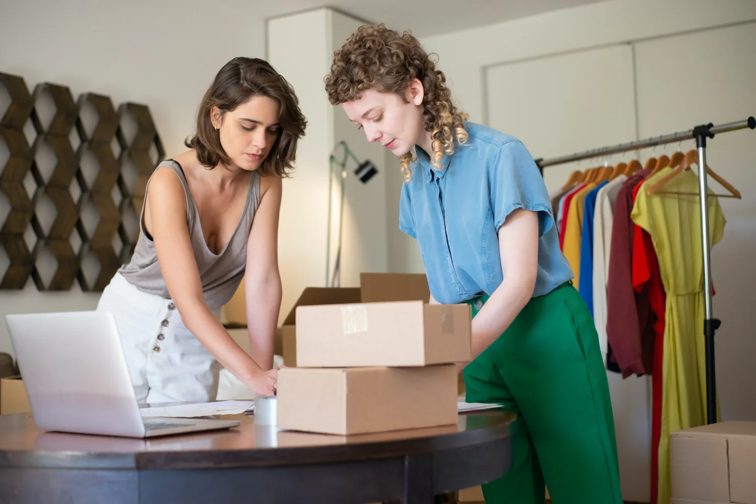 Two people pack boxes for shipping on a wooden table with a laptop open